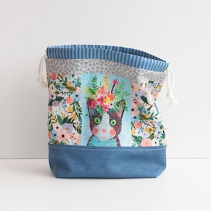 May include: A drawstring bag with a cat illustration wearing a floral crown. The bag features a blue denim base, a floral pattern, and a striped blue top. White drawstrings are visible.