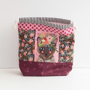 Puede incluir: Una bolsa con cordón con una ilustración de gato. La bolsa presenta un estampado floral, lunares rosas y una parte superior a rayas. La ilustración del gato está en el centro, con una corona de flores. La parte inferior de la bolsa es de color burdeos liso.