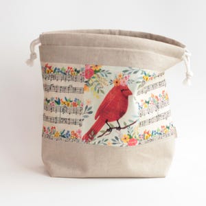 May include: A beige drawstring pouch featuring a central panel with a red cardinal bird wearing a floral crown, surrounded by musical notes and colorful flowers. The bag is made of a light-colored fabric with white cord ties at the top opening.