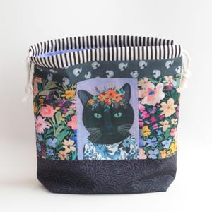 Puede incluir: Una bolsa de tela con cordón que presenta un panel central con un gato negro con una corona de flores, flanqueado por coloridos patrones florales sobre un fondo oscuro. El borde superior tiene una franja a rayas blancas y negras y cordones.