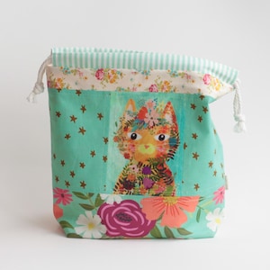 Puede incluir: Una bolsa de tela con cordón que presenta un fondo verde azulado con estrellas doradas, una ilustración central de un gato adornado con flores coloridas y un borde inferior con grandes patrones florales en rosa, naranja y blanco.