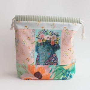 Peut inclure: Une pochette en tissu à cordon de serrage avec un panneau central représentant un éléphant bleu portant une couronne de fleurs. Le sac présente des panneaux latéraux à motifs floraux roses, une base à grandes fleurs orange et vertes, et un haut rayé vert sauge.