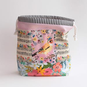 May include: A fabric drawstring pouch featuring a central panel with a yellow bird and floral print, flanked by sections of musical notation and colorful flowers. The top edge has a black and white striped trim with white cotton drawstrings.