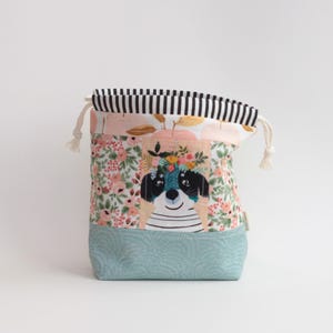 May include: A fabric drawstring pouch featuring a central illustration of a dog wearing a floral crown, surrounded by pink and green floral patterns. The base is a solid light blue with a fan-like texture, and the top has black and white stripes.