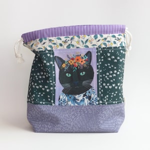 May include: A fabric drawstring bag featuring a central panel with a black cat wearing a floral crown. The bag includes sections of dark green fabric with white floral patterns, a top band of light purple stripes, and a light purple base with white dashes.