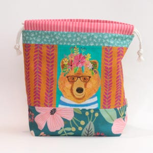 May include: A fabric drawstring pouch featuring a central illustration of a brown bear wearing glasses and a floral crown. The bag has a teal floral base, orange side panels with a leaf pattern, and a pink and white striped top with white cords.