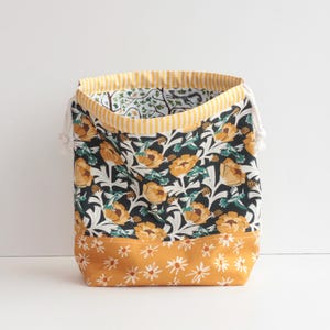 May include: A fabric storage bag with a floral pattern. The bag features a black background with yellow flowers and green leaves. The top edge has a yellow and white striped pattern, and the bottom has a yellow floral pattern. White drawstrings are visible.