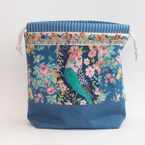 May include: A drawstring project bag featuring a blue fabric base with a wave pattern, a central panel with a budgie bird and floral print, and a top section with blue and white vertical stripes. The bag includes white cord ties and musical notes.