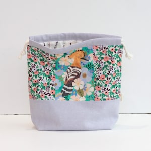 May include: A fabric drawstring bag with a floral and bird design. The bag features a light gray base and a top section with a colorful pattern of flowers and a hoopoe bird. The drawstring is white.