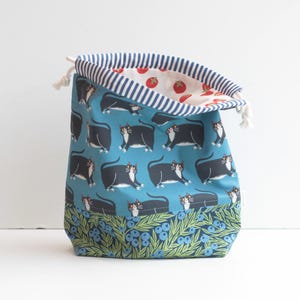 May include: A drawstring bag with a teal background featuring black and white cat illustrations. The bag's interior has a striped pattern with red tomato illustrations. The bottom border has a floral design with blueberries.