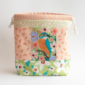 May include: A fabric drawstring bag featuring a central panel with a kingfisher bird surrounded by flowers. The bag includes side panels with a small shrimp pattern, a green base with white floral motifs, and a top section with orange stripes.