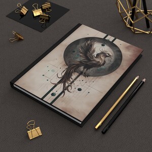 May include: A black journal with a phoenix illustration on the cover. The artwork features a detailed phoenix with brown and white feathers set against a dark gray circle and geometric lines. Gold and black pencils and gold binder clips are also present.