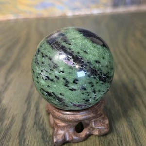 Pode incluir: Uma pedra esf&eacute;rica polida com uma base verde vibrante, estrias pretas proeminentes e pequenas manchas de magenta profundo. A esfera repousa sobre um suporte de madeira esculpida em castanho escuro com base circular e tr&ecirc;s pernas curvas.