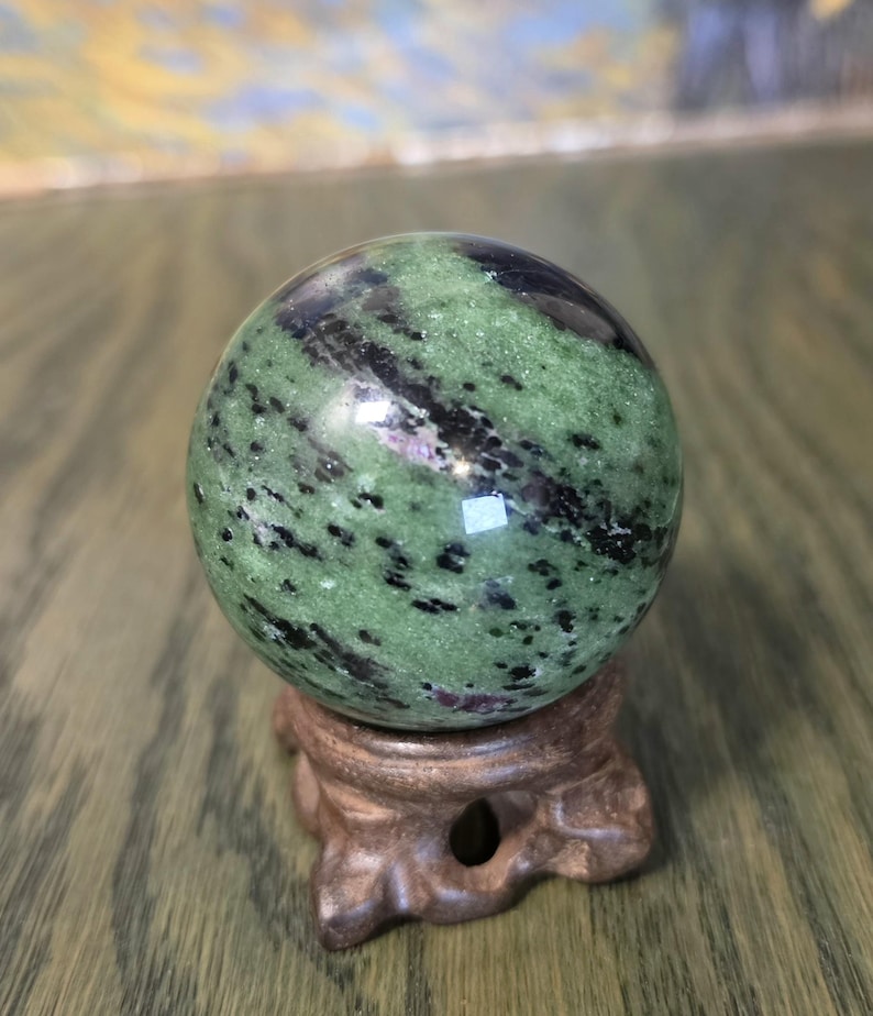 Pode incluir: Uma pedra esf&eacute;rica polida com uma base verde vibrante, estrias pretas proeminentes e pequenas manchas de magenta profundo. A esfera repousa sobre um suporte de madeira esculpida em castanho escuro com base circular e tr&ecirc;s pernas curvas.