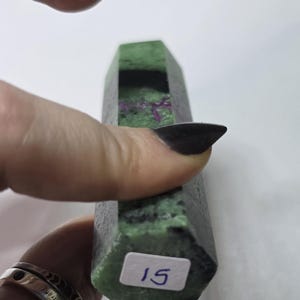 May include: A hexagonal mineral specimen featuring a mottled green and black surface with embedded magenta inclusions. A white rectangular sticker with the handwritten number 15 is affixed to the flat base of the stone, which has a polished finish.