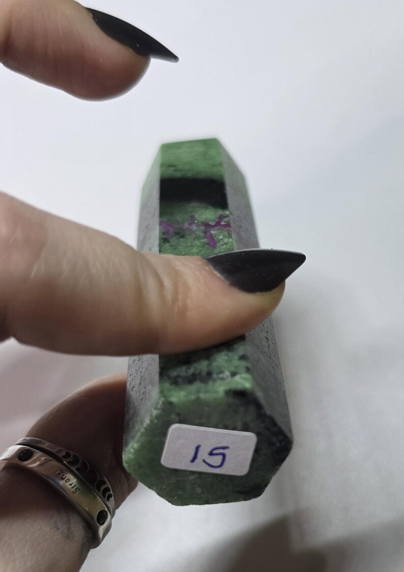 May include: A hexagonal mineral specimen featuring a mottled green and black surface with embedded magenta inclusions. A white rectangular sticker with the handwritten number 15 is affixed to the flat base of the stone, which has a polished finish.