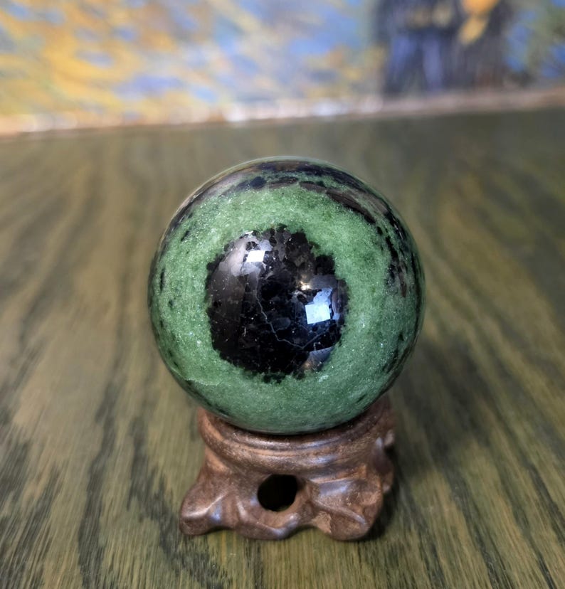 Pode incluir: Uma pedra esf&eacute;rica polida com uma base verde vibrante e uma inclus&atilde;o circular preta proeminente no centro. A esfera &eacute; apoiada por um pequeno suporte de madeira castanha esculpida com um design de base decorativo e aberto.