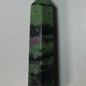 May include: A polished zoisite crystal tower with a hexagonal shape, featuring a vibrant green base colour, prominent black mineral inclusions in horizontal bands, and a distinct patch of deep magenta ruby visible on the front surface.