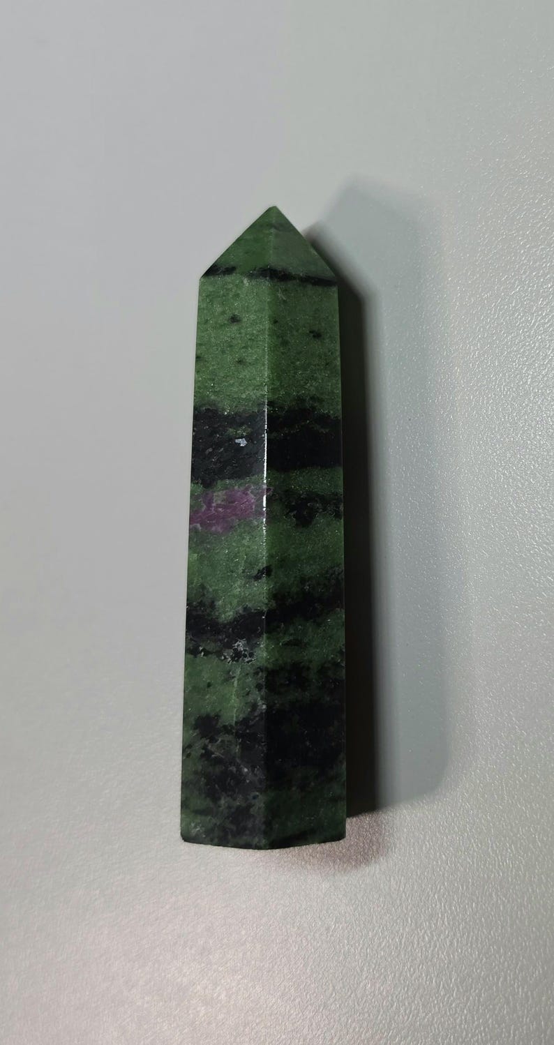 May include: A polished zoisite crystal tower with a hexagonal shape, featuring a vibrant green base colour, prominent black mineral inclusions in horizontal bands, and a distinct patch of deep magenta ruby visible on the front surface.