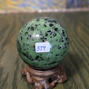 Pode incluir: Uma esfera de pedra polida com manchas verdes e pretas, apoiada num suporte de madeira castanha esculpida. Um pequeno autocolante retangular branco com o n&uacute;mero 379 escrito &agrave; m&atilde;o em azul est&aacute; colado no centro da superf&iacute;cie mineral.