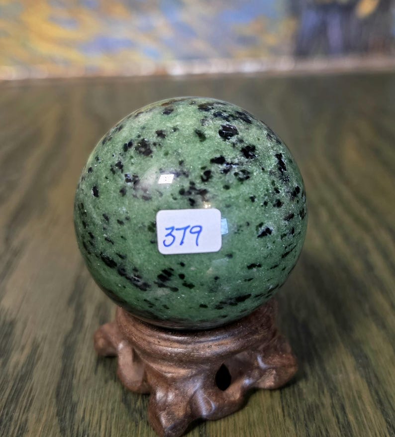 Pode incluir: Uma esfera de pedra polida com manchas verdes e pretas, apoiada num suporte de madeira castanha esculpida. Um pequeno autocolante retangular branco com o n&uacute;mero 379 escrito &agrave; m&atilde;o em azul est&aacute; colado no centro da superf&iacute;cie mineral.