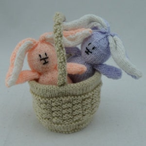 May include: A knitted basket contains two small bunny toys. One bunny is peach with white-tipped ears, the other is lavender with white-tipped ears. The basket and handle are beige. The bunnies have black stitched eyes and mouths.