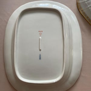 May include: White oval serving platter with a slightly raised edge. The underside features a red and black stamp with the letters "KPM" and a stylized symbol. A small blue mark is also visible.