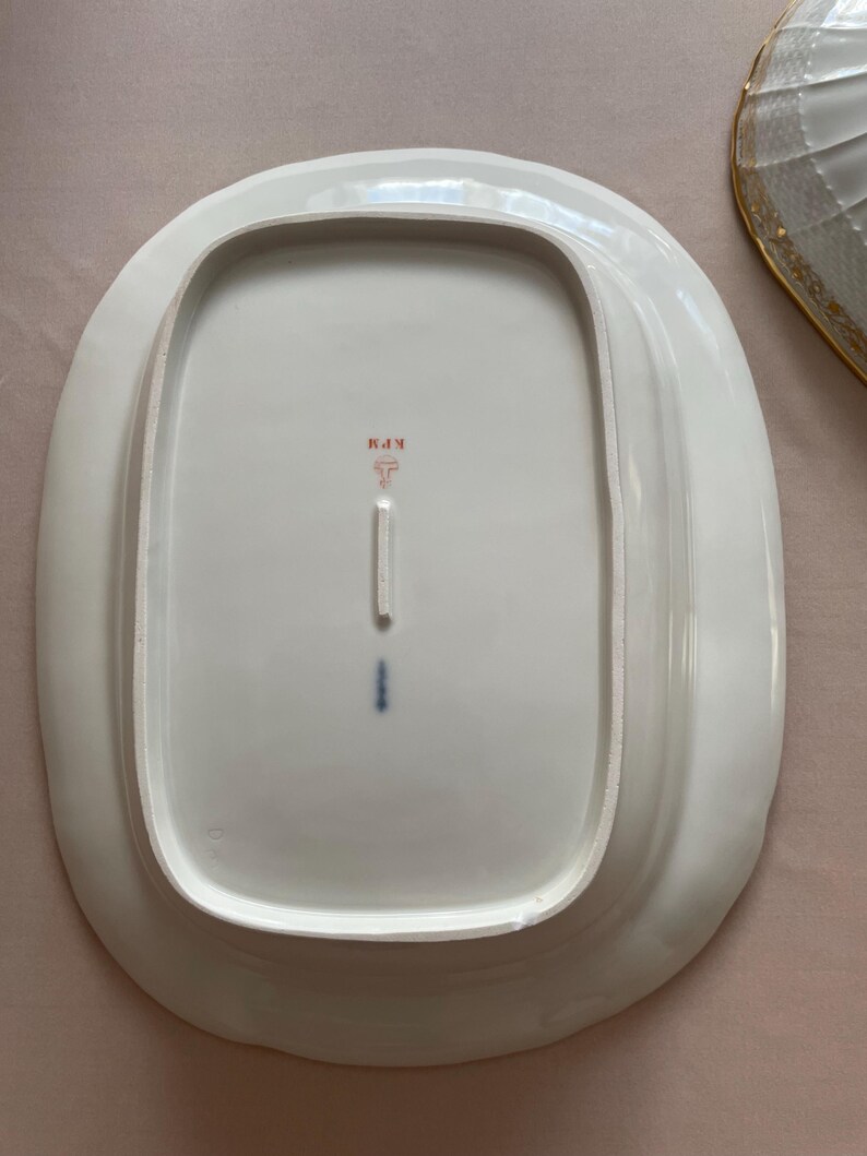 May include: White oval serving platter with a slightly raised edge. The underside features a red and black stamp with the letters "KPM" and a stylized symbol. A small blue mark is also visible.