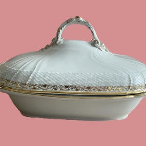 May include: White porcelain serving dish with a lid and gold trim. The dish has a decorative handle and a textured pattern on the lid. The rim features a gold floral design. A classic piece for serving food.