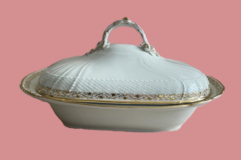 May include: White porcelain serving dish with a lid and gold trim. The dish has a decorative handle and a textured pattern on the lid. The rim features a gold floral design. A classic piece for serving food.