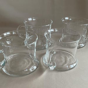 May include: Four clear glass mugs with rounded handles and wide bases. The mugs have a classic design, suitable for serving hot beverages. The transparent glass allows the contents to be visible. The mugs are arranged on a neutral-toned surface.