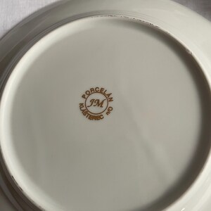 May include: Off-white porcelain plate with a gold rim. The underside features a gold emblem with the text "PORCELAN JM KLASTEREC n/o". The plate has a slightly curved edge.