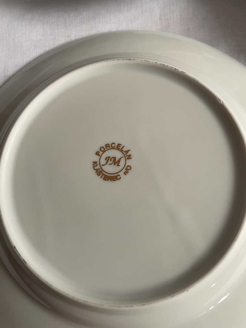 May include: Off-white porcelain plate with a gold rim. The underside features a gold emblem with the text "PORCELAN JM KLASTEREC n/o". The plate has a slightly curved edge.