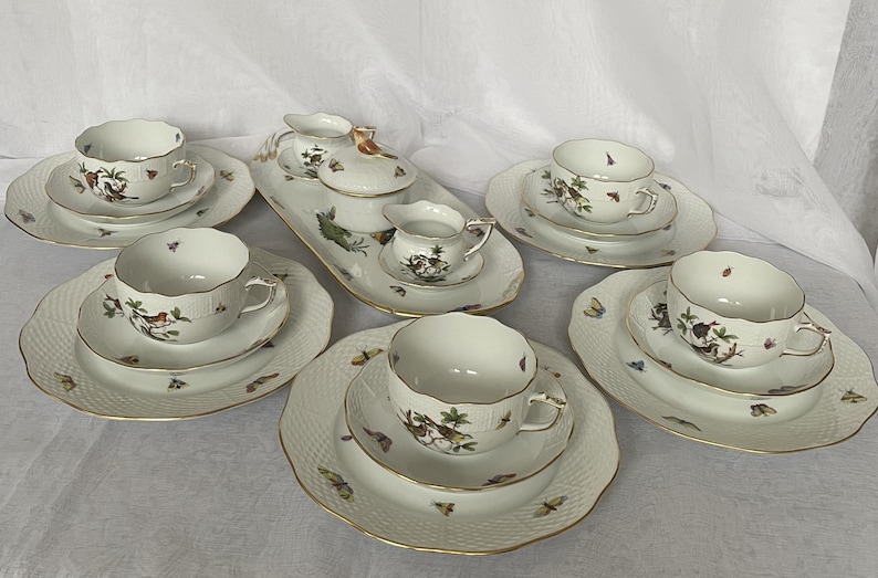 May include: A white porcelain tea set with gold trim, featuring cups, saucers, and serving pieces. The set is decorated with bird and butterfly motifs. The plates have a textured pattern.