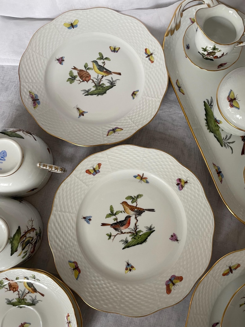 May include: A collection of white porcelain dishes with a textured rim and gold trim. Each piece features a detailed illustration of birds perched on branches, surrounded by colorful butterflies. Includes plates, bowls, and serving pieces.