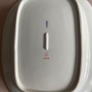 May include: A white, oval porcelain platter with a raised rim. The underside has a blue vertical mark, a white rectangular mark, and a red stamp with the letters "KPM". The platter has a smooth, glossy finish.