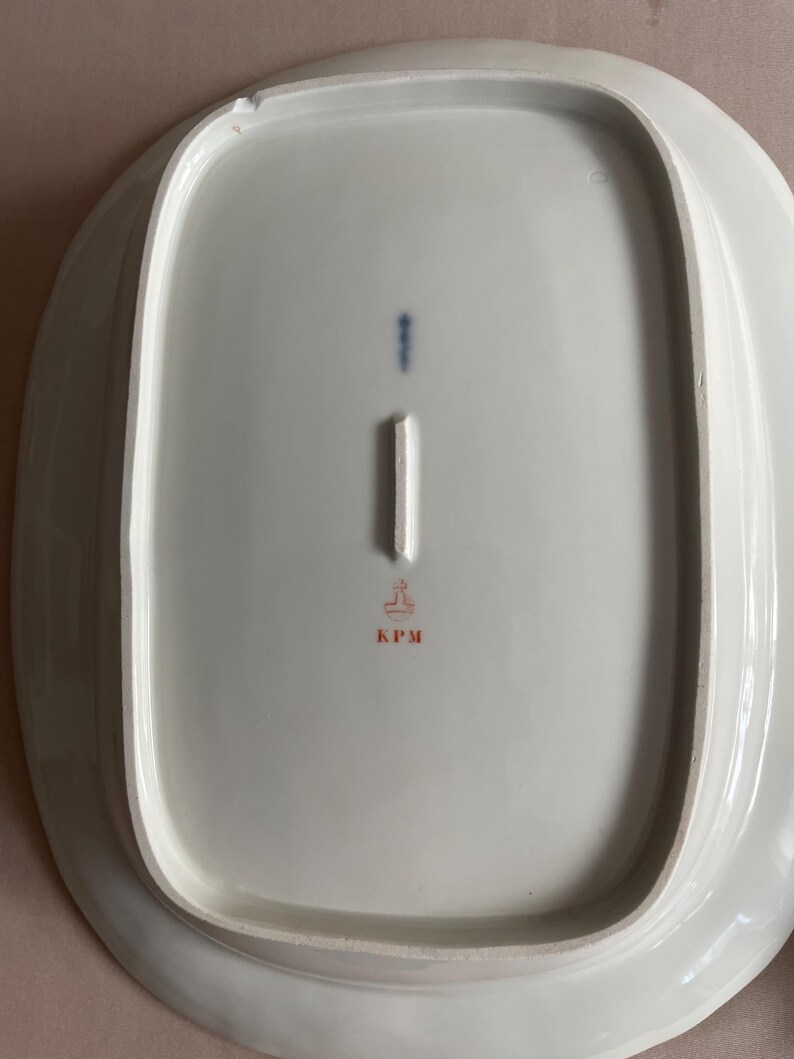May include: A white, oval porcelain platter with a raised rim. The underside has a blue vertical mark, a white rectangular mark, and a red stamp with the letters "KPM". The platter has a smooth, glossy finish.