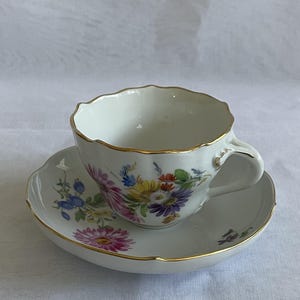 May include: White teacup and saucer set with a floral design in pink, purple, yellow, and blue. The cup and saucer have a gold trim. The cup has a scalloped rim and a curved handle. The saucer has a matching floral pattern.