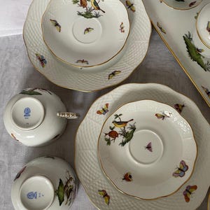 May include: A collection of white porcelain tableware with gold trim, featuring bird and butterfly designs. The set includes plates, saucers, and teacups. The plates have a textured surface. The teacups have decorative handles.