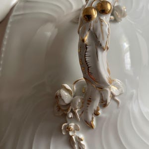 May include: Close-up of a white porcelain serving dish lid with a decorative handle. The handle has gold accents and floral details, including two golden spheres. The dish has a ridged design.