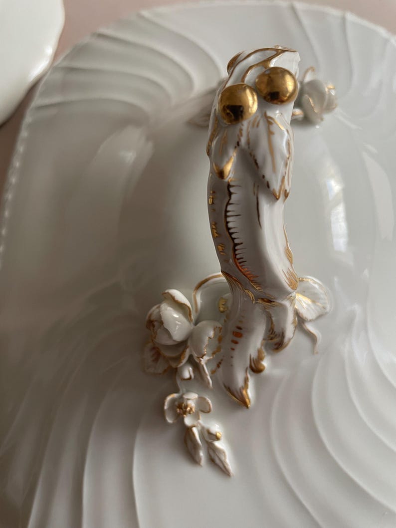 May include: Close-up of a white porcelain serving dish lid with a decorative handle. The handle has gold accents and floral details, including two golden spheres. The dish has a ridged design.