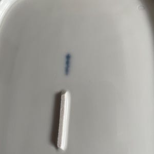 May include: A close-up of a white porcelain dish with a rectangular indentation and a blue mark. The dish has a slightly curved edge and a small, faded red emblem at the bottom.