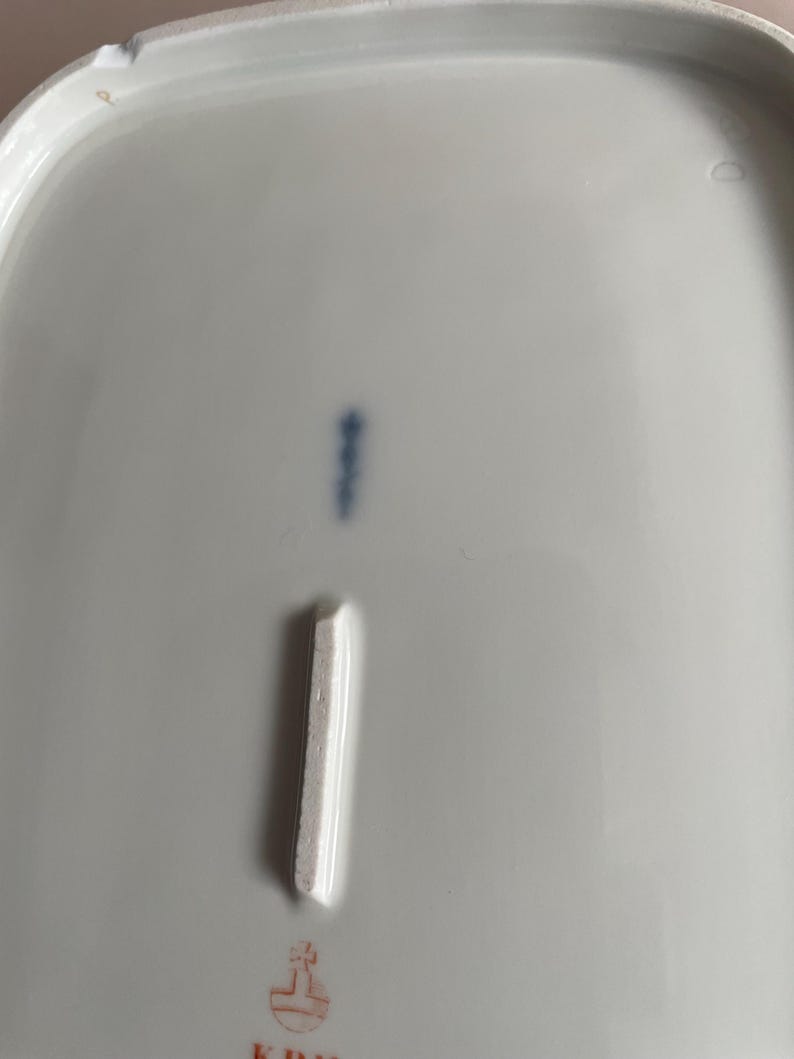 May include: A close-up of a white porcelain dish with a rectangular indentation and a blue mark. The dish has a slightly curved edge and a small, faded red emblem at the bottom.