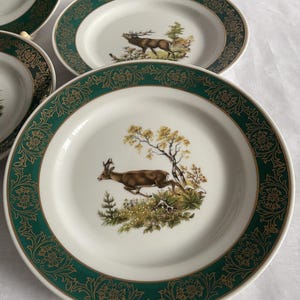 May include: A set of white ceramic plates with a green and gold floral border. Each plate features a detailed illustration of a deer in a natural landscape. The plates are designed for serving food.