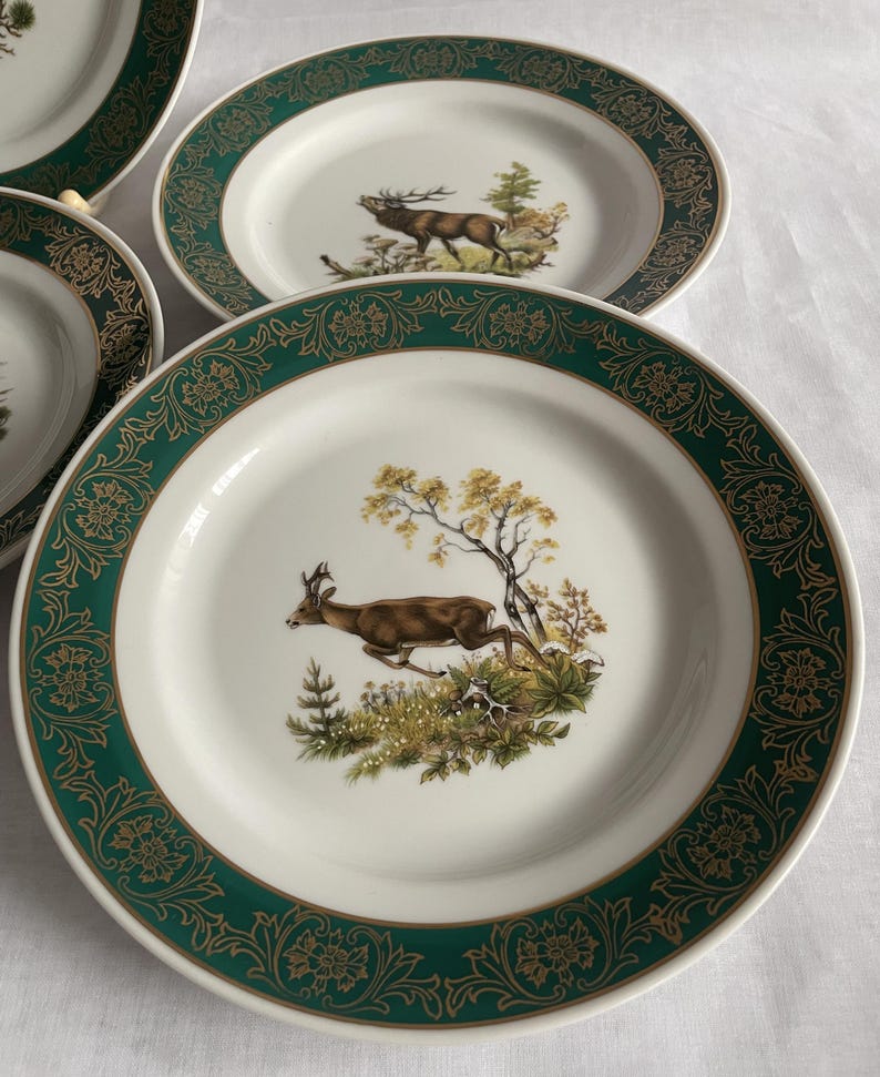 May include: A set of white ceramic plates with a green and gold floral border. Each plate features a detailed illustration of a deer in a natural landscape. The plates are designed for serving food.