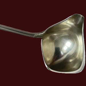 May include: A silver gravy boat ladle with a long, curved handle and a deep, rounded bowl. The ladle is made of shiny metal and reflects the surrounding light. The handle has a ridged design for grip.