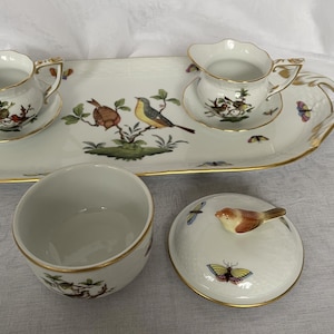 May include: A white porcelain tea set with gold trim, featuring bird and butterfly illustrations. Includes a serving tray, two teacups, a creamer, and a covered bowl. The lid of the bowl has a bird-shaped handle.