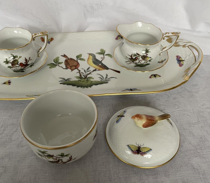 May include: A white porcelain tea set with gold trim, featuring bird and butterfly illustrations. Includes a serving tray, two teacups, a creamer, and a covered bowl. The lid of the bowl has a bird-shaped handle.