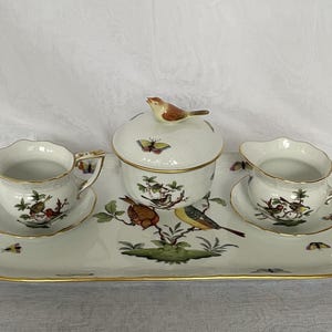 May include: A porcelain tea set with a serving tray, sugar bowl, and two cream pitchers. The set is decorated with colorful birds, butterflies, and floral designs, with gold trim. The tray measures approximately 16 inches long.