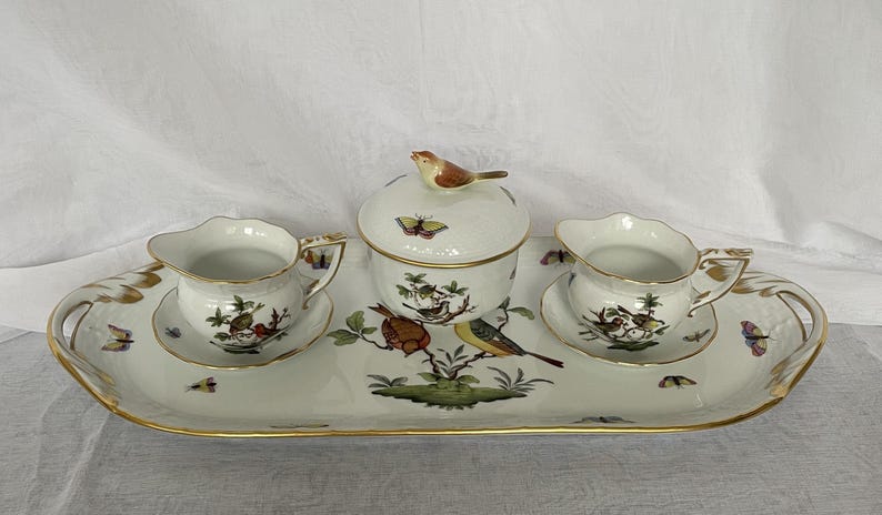 May include: A porcelain tea set with a serving tray, sugar bowl, and two cream pitchers. The set is decorated with colorful birds, butterflies, and floral designs, with gold trim. The tray measures approximately 16 inches long.